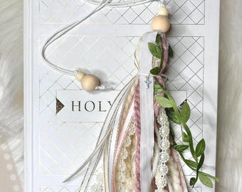 May include: A white book with the word "HOLY" printed on the cover. A pink, white, and gold tassel bookmark with a small silver cross charm is attached to the book.
