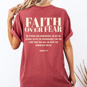 May include: A maroon t-shirt with the words "FAITH OVER FEAR" printed across the back in a cream-colored font. Below the text is a quote from Joshua 1:9. The shirt has rolled-up sleeves.