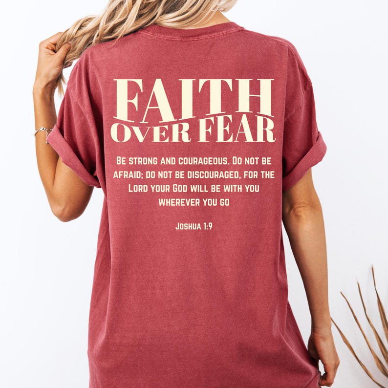 May include: A maroon t-shirt with the words "FAITH OVER FEAR" printed across the back in a cream-colored font. Below the text is a quote from Joshua 1:9. The shirt has rolled-up sleeves.