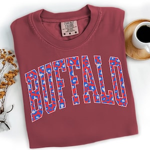 May include: A maroon Comfort Colors t-shirt with the word "BUFFALO" in a white outline, filled with a blue and red leopard print design. The shirt is folded, with a tag visible at the neckline. A cup of coffee sits on a wooden coaster.