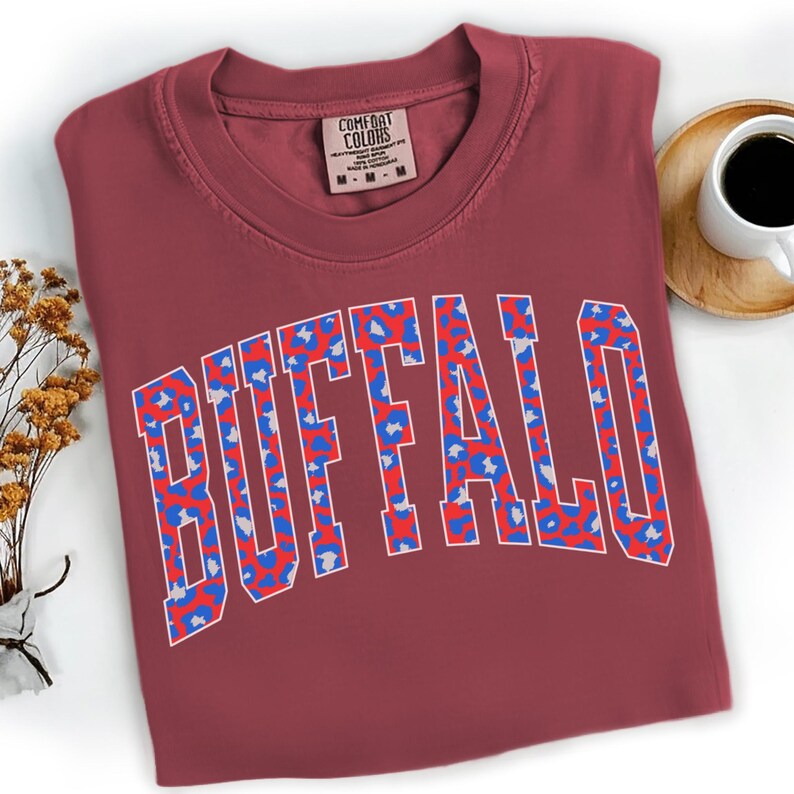 May include: A maroon Comfort Colors t-shirt with the word "BUFFALO" in a white outline, filled with a blue and red leopard print design. The shirt is folded, with a tag visible at the neckline. A cup of coffee sits on a wooden coaster.