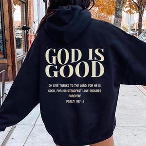 Könnte beinhalten: Schwarzer Kapuzenpullover mit dem Text "GOD IS GOOD" in weißen Buchstaben. Der Text "OH GIVE THANKS TO THE LORD, FOR HE IS GOOD, FOR HIS STEADFAST LOVE ENDURES FOREVER! PSALM 107:1" ist darunter in kleineren weißen Buchstaben gedruckt.