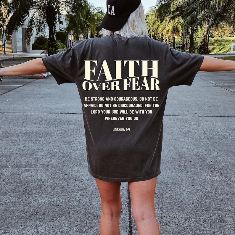 May include: Dark gray t-shirt with the text "FAITH OVER FEAR" in white block letters. Below the text is a quote from Joshua 1:9. The shirt is a short-sleeved, casual style.