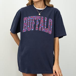 May include: Navy blue t-shirt with the word "BUFFALO" in a red and blue floral pattern across the chest. The shirt has short sleeves and a crew neck. The model is wearing a white undershirt and a gold necklace.
