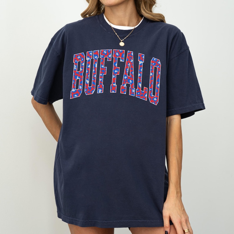 May include: Navy blue t-shirt with the word "BUFFALO" in a red and blue floral pattern across the chest. The shirt has short sleeves and a crew neck. The model is wearing a white undershirt and a gold necklace.