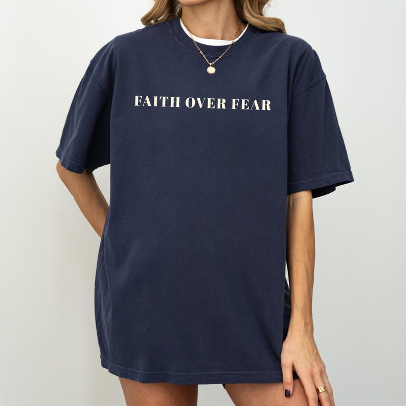 May include: Navy blue t-shirt with the text "FAITH OVER FEAR" in white block letters. The shirt has short sleeves and a relaxed fit. The model is wearing a white undershirt and a gold necklace.