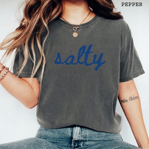 以下が含まれることがあります： 青い文字で「salty」と書かれた濃い灰色のTシャツ。Tシャツはクルーネックで半袖です。「pepper」という文字がTシャツの右上隅に印刷されています。