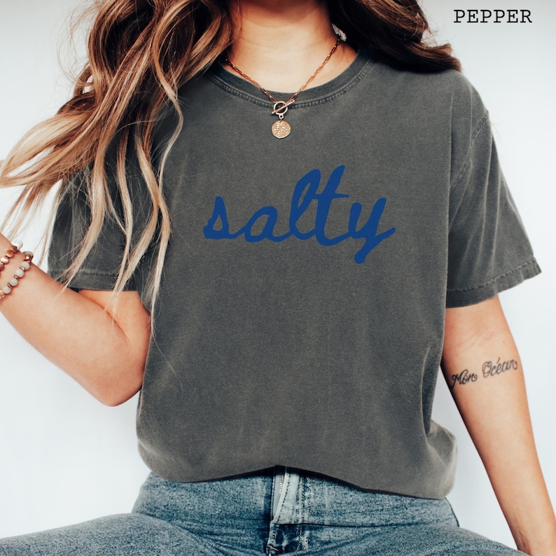 以下が含まれることがあります： 青い文字で「salty」と書かれた濃い灰色のTシャツ。Tシャツはクルーネックで半袖です。「pepper」という文字がTシャツの右上隅に印刷されています。