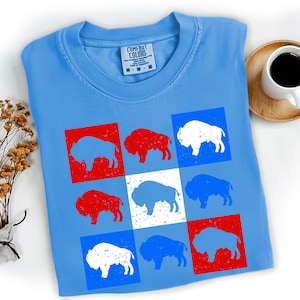 May include: Light blue t-shirt featuring a grid of nine squares, each containing a buffalo silhouette. The buffaloes are in red, white, and blue, with a Comfort Colors tag at the neckline. A coffee cup and decorative plant are in the background.