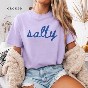以下が含まれることがあります： 水色の文字で「salty」と書かれたライラック色のTシャツ。「ORCHID」の文字が左上にプリントされています。半袖のシャツは柔らかい素材で作られているようです。