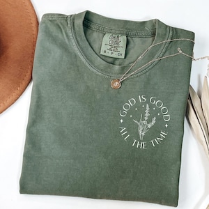 May include: Olive green Comfort Colors t-shirt with the phrase "GOD IS GOOD ALL THE TIME" in a white circular design. A delicate gold necklace with a pendant rests on the shirt. The shirt is folded.
