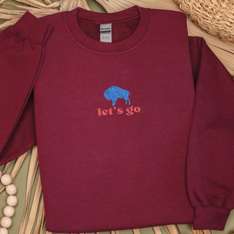 May include: Burgundy crewneck sweatshirt with a blue glitter buffalo graphic and the text "let's go" in red. The sweatshirt is made of a soft material and has long sleeves. The Gildan tag is visible.