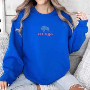 May include: Royal blue sweatshirt with a buffalo graphic and the text "let's go" in red. The buffalo graphic is a detailed outline. The sweatshirt has long sleeves and a crew neck.