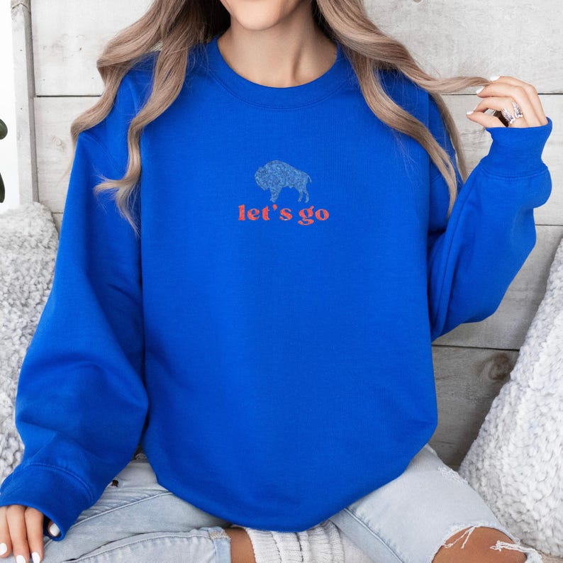 May include: Royal blue sweatshirt with a buffalo graphic and the text "let's go" in red. The buffalo graphic is a detailed outline. The sweatshirt has long sleeves and a crew neck.