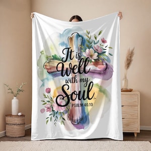 May include: A white blanket with a watercolour cross design. The text reads "It is Well with my Soul" and "Psalm 46:10". The cross is decorated with pink, green, and purple floral accents, set against a pastel-coloured background.