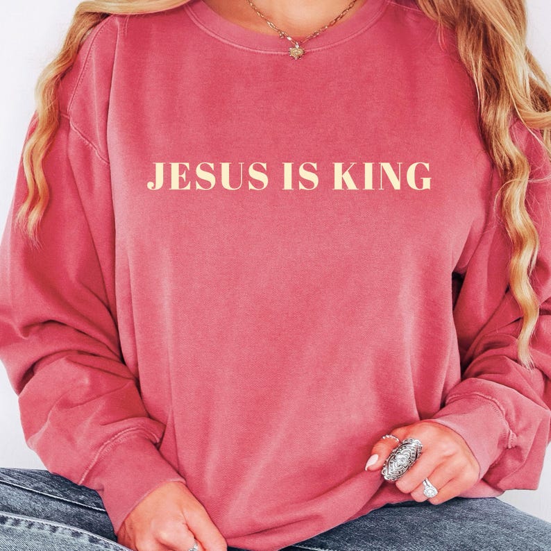 May include: A dusty rose-colored sweatshirt with the text "JESUS IS KING" in a light beige font. The long-sleeved top is worn by a person, with their hands and jewelry visible. The overall style is casual and comfortable.