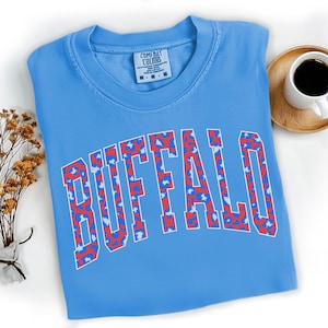 May include: A light blue t-shirt with the word "BUFFALO" in a white, red, and blue leopard print design. The shirt has a crew neck and short sleeves. A small tag with the brand name "COMFORT COLORS" is visible on the shirt.