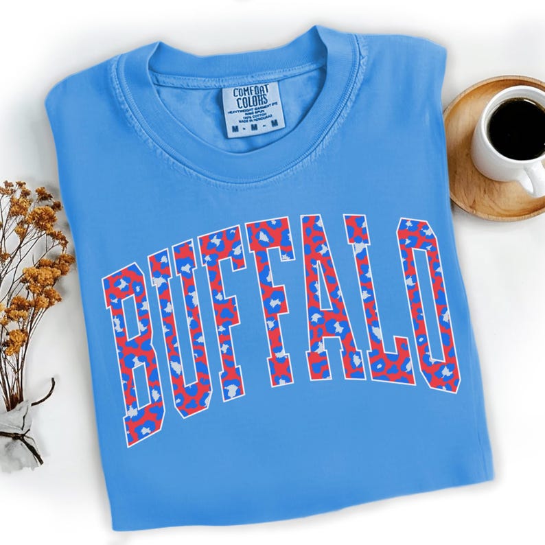 May include: A light blue t-shirt with the word "BUFFALO" in a white, red, and blue leopard print design. The shirt has a crew neck and short sleeves. A small tag with the brand name "COMFORT COLORS" is visible on the shirt.