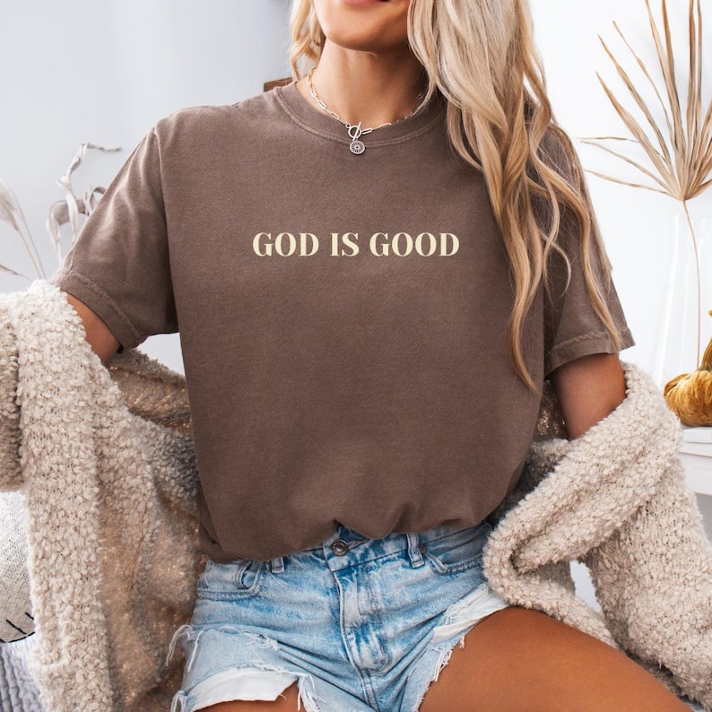 May include: A brown t-shirt with the text "GOD IS GOOD" printed in white on the front. The shirt is being worn by a person with long blonde hair and is paired with denim shorts.