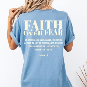 May include: A light blue t-shirt with the text "FAITH OVER FEAR" in a cream-colored font. Below the text is a quote from Joshua 1:9. The shirt has rolled-up sleeves.