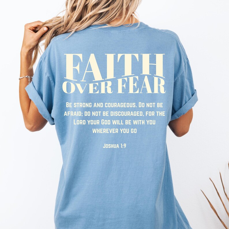 May include: A light blue t-shirt with the text "FAITH OVER FEAR" in a cream-colored font. Below the text is a quote from Joshua 1:9. The shirt has rolled-up sleeves.