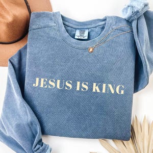 May include: A blue crewneck sweatshirt with the text "JESUS IS KING" in a light tan color. A gold necklace with a pendant is draped on the sweatshirt. A brown hat is visible in the background.