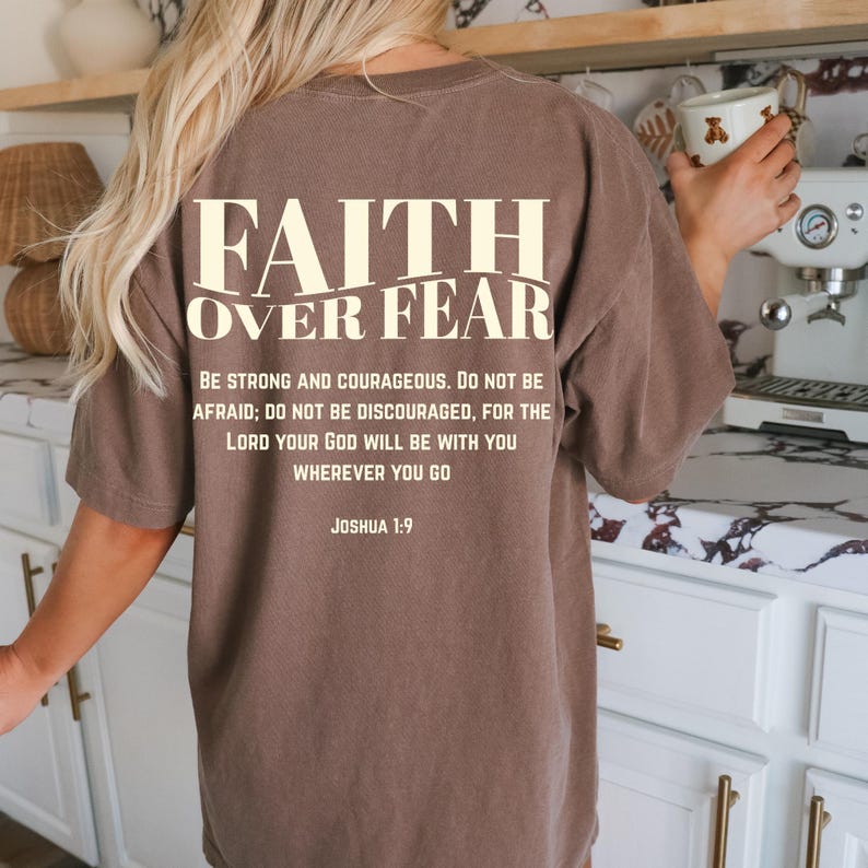 May include: A brown t-shirt with the words "FAITH OVER FEAR" printed on the back in a cream color. Below the text is a quote from Joshua 1:9. The shirt is a solid color and has a relaxed fit.
