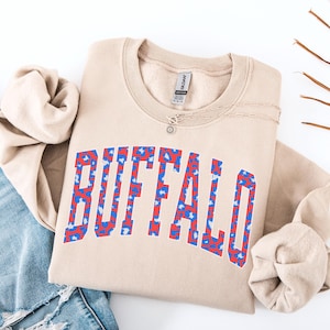 May include: A tan sweatshirt with the word "BUFFALO" in large, bold letters across the front. The letters are filled with a red and blue pattern. A silver necklace with a sun-shaped pendant is on the sweatshirt.