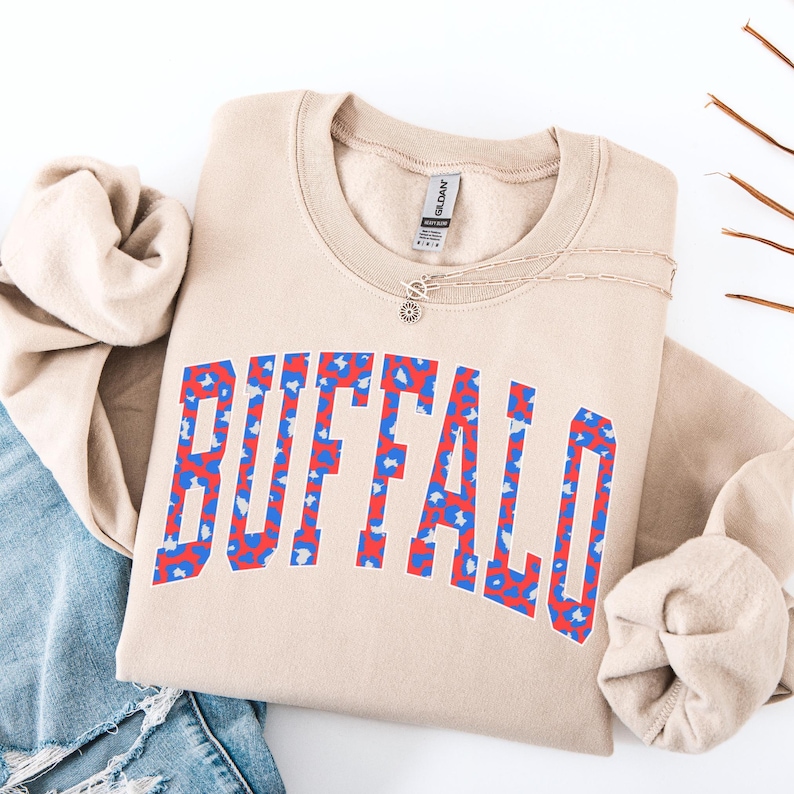 May include: A tan sweatshirt with the word "BUFFALO" in large, bold letters across the front. The letters are filled with a red and blue pattern. A silver necklace with a sun-shaped pendant is on the sweatshirt.