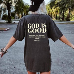 May include: A dark gray t-shirt with the text "GOD IS GOOD" in white lettering. The shirt also includes the text "ON GIVE THANKS TO THE LORD, FOR HE IS GOOD, FOR HIS STEADFAST LOVE ENDURES FOREVER! PSALM 107:1" in smaller white lettering.