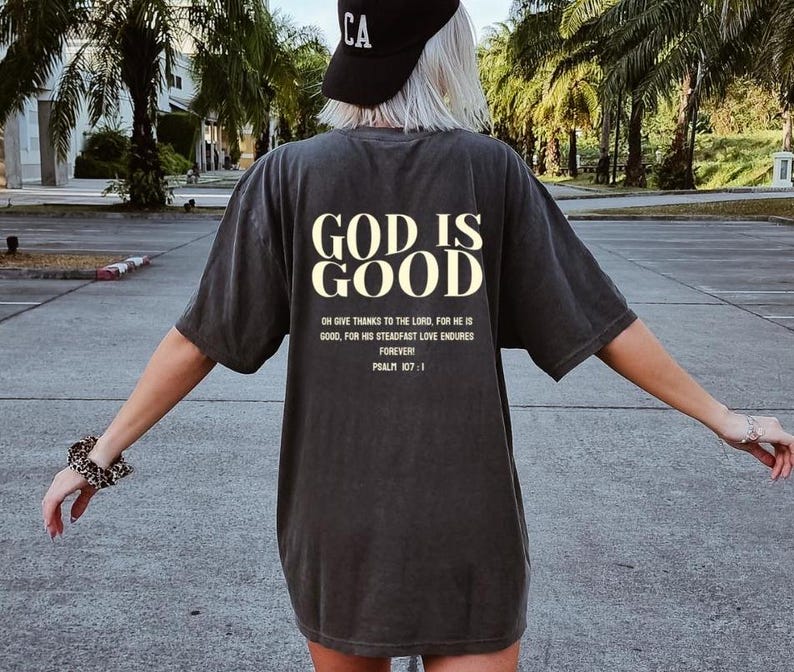May include: A dark gray t-shirt with the text "GOD IS GOOD" in white lettering. The shirt also includes the text "ON GIVE THANKS TO THE LORD, FOR HE IS GOOD, FOR HIS STEADFAST LOVE ENDURES FOREVER! PSALM 107:1" in smaller white lettering.