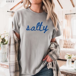 Grey unisex graphic t-shirt featuring the word “salty” in bold blue handwritten script across the chest. Soft neutral tee with a relaxed fit, styled casually with jeans and a plaid layer for a cozy everyday look. Minimalist statement design with a coastal, beach-inspired vibe, perfect for summer outfits, casual wear, or ocean lovers who enjoy fun graphic tees.