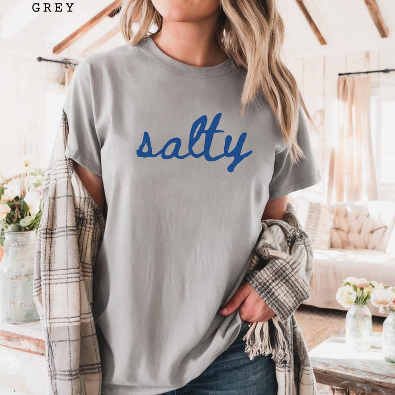 Grey unisex graphic t-shirt featuring the word “salty” in bold blue handwritten script across the chest. Soft neutral tee with a relaxed fit, styled casually with jeans and a plaid layer for a cozy everyday look. Minimalist statement design with a coastal, beach-inspired vibe, perfect for summer outfits, casual wear, or ocean lovers who enjoy fun graphic tees.