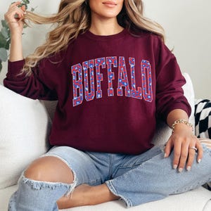 May include: Burgundy sweatshirt with the word "BUFFALO" in large, block letters across the chest. The letters are filled with a blue and red pattern. The sweatshirt has long sleeves and a crew neck.
