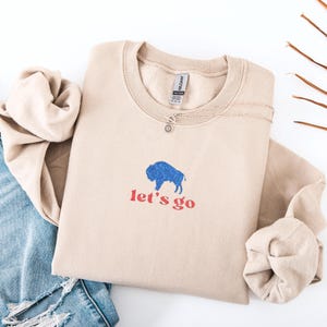 May include: A beige sweatshirt with a blue bison graphic and the words "let's go" in red. A silver necklace with a small pendant is on the sweatshirt. The sweatshirt is folded next to a pair of blue jeans.