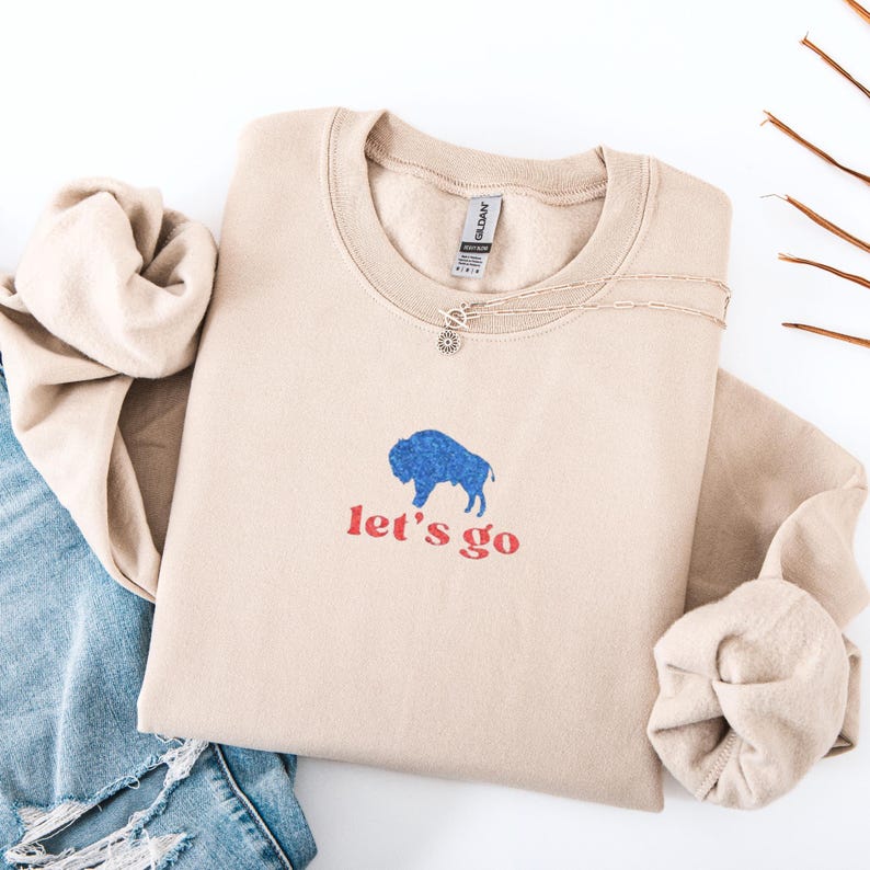 May include: A beige sweatshirt with a blue bison graphic and the words "let's go" in red. A silver necklace with a small pendant is on the sweatshirt. The sweatshirt is folded next to a pair of blue jeans.