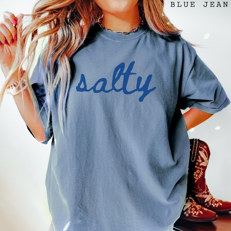 Blue Jean unisex graphic t-shirt featuring the word “salty” in bold navy handwritten script across the chest. Relaxed fit tee with a coastal, beach-inspired vibe, styled casually with layered jewelry and western boots for a trendy everyday look. Soft washed blue color makes it perfect for summer outfits, vacation wear, ocean lovers, and fun statement style.