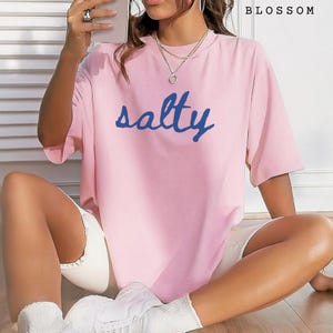 Blossom pink unisex graphic t-shirt featuring the word “salty” in bold blue handwritten script across the chest. Soft pastel tee with a relaxed oversized fit, styled casually with white shorts, sneakers, and layered jewelry. Trendy coastal-inspired statement shirt perfect for summer outfits, beach days, vacation wear, lounging, or ocean lovers who enjoy fun graphic tees.