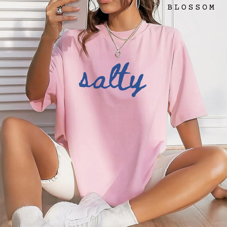 Blossom pink unisex graphic t-shirt featuring the word “salty” in bold blue handwritten script across the chest. Soft pastel tee with a relaxed oversized fit, styled casually with white shorts, sneakers, and layered jewelry. Trendy coastal-inspired statement shirt perfect for summer outfits, beach days, vacation wear, lounging, or ocean lovers who enjoy fun graphic tees.