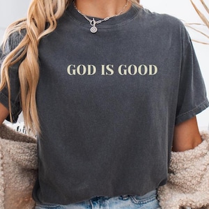 May include: A dark gray cropped t-shirt with the text "GOD IS GOOD" in white lettering.