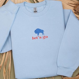 May include: A light blue sweatshirt with a blue buffalo graphic and the words "let's go" in red. The sweatshirt is folded, showing the sleeve and part of the collar. The background is a neutral color.