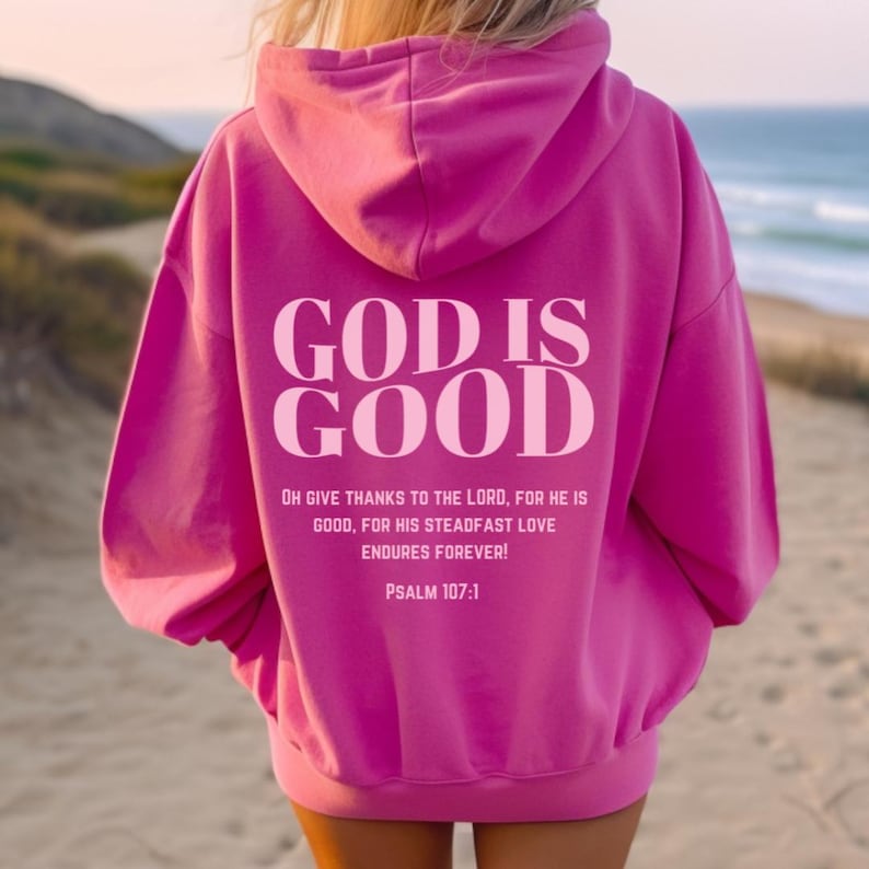 May include: Pink hoodie with the text "GOD IS GOOD" in white lettering. The text "OH GIVE THANKS TO THE LORD, FOR HE IS GOOD, FOR HIS STEADFAST LOVE ENDURES FOREVER! PSALM 107:1" is printed below the main text.