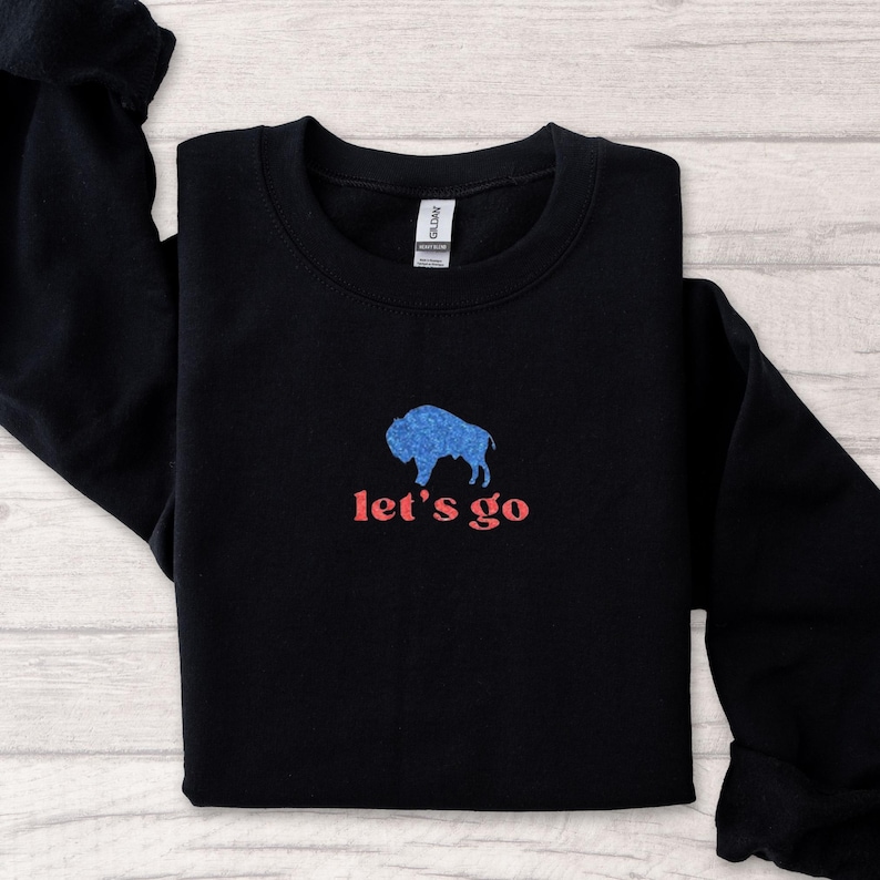 May include: Black sweatshirt with a blue buffalo graphic and the text "let's go" in red. The sweatshirt is folded, showcasing the design on the front. The design features a textured buffalo silhouette.