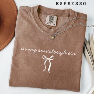 May include: Espresso-colored t-shirt with the phrase "in my sourdough era" and a bow graphic in white. The shirt has a crew neck and short sleeves. A silver necklace with a pendant is draped across the shirt.