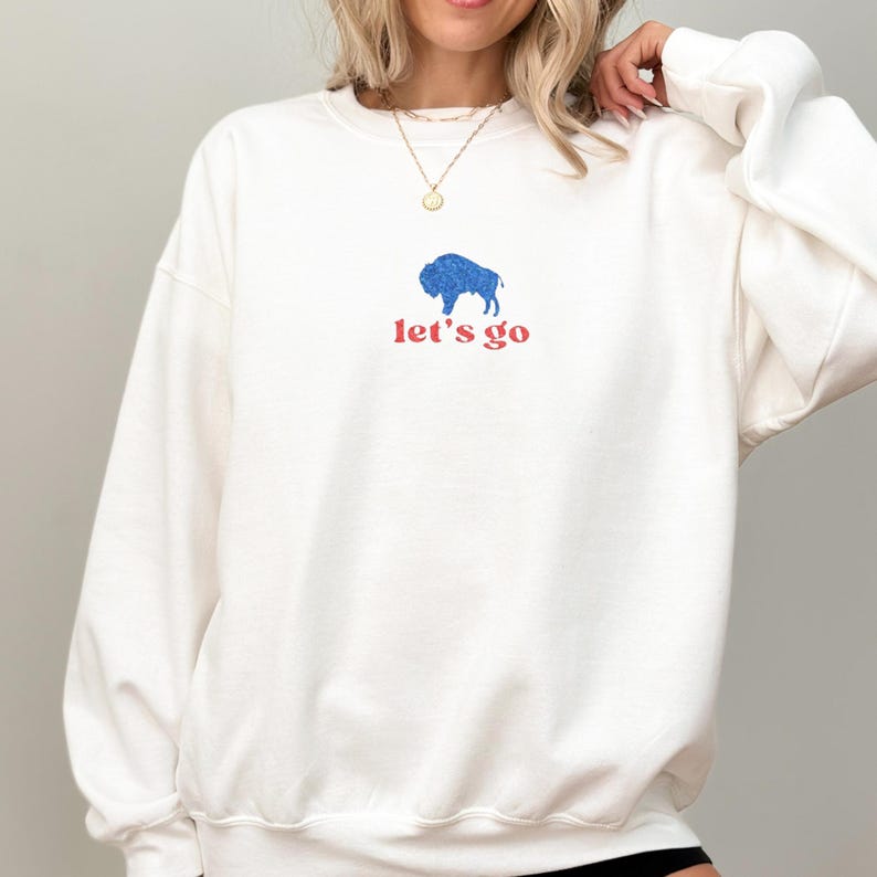 May include: Cream-colored sweatshirt with a blue buffalo graphic and the words "let's go" in red. The design is centered on the front of the long-sleeved crew neck top. The sweatshirt is made of a soft material.
