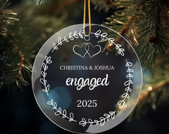 May include: Clear, round Christmas ornament with the words "Christina & Joshua engaged 2025." The ornament has a white floral design around the edge and two intertwined hearts. It hangs from a gold ribbon.