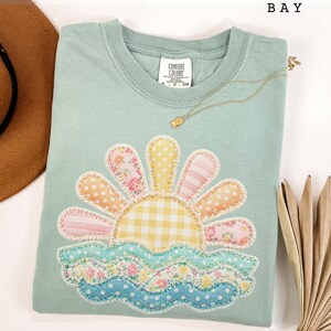 Op de afbeelding: Een lichtgroen Comfort Colors T-shirt met een kleurrijk zon- en golfontwerp. De zon heeft bloemen-, stippen- en strepenpatronen. Een gouden ketting is over het shirt gedrapeerd. Het woord "BAY" staat bovenaan gedrukt.