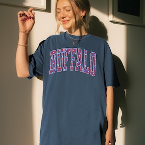 May include: A navy blue t-shirt with the word "BUFFALO" in a red, white, and blue patterned design. The shirt has short sleeves and a relaxed fit. The t-shirt is a casual wear item.