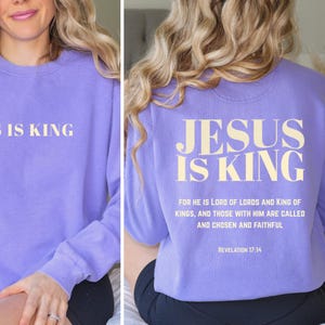 May include: A lilac-colored sweatshirt with the text "JESUS IS KING" printed on the front and back. The back of the sweatshirt also features a Bible verse from Revelation 17:14.