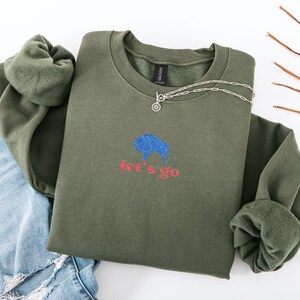 May include: An olive green sweatshirt with a blue bison graphic and the words "let's go" in red. A silver necklace with a circular pendant is on the sweatshirt. The sweatshirt is folded with a pair of blue jeans.
